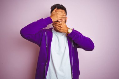 Mor eşofman üstü giyen genç Hintli adam izole edilmiş pembe arka plan üzerinde duruyor gözlerini ve ağzını elleriyle kapıyor, şaşırmış ve şok olmuş. Duygularını gizliyor.