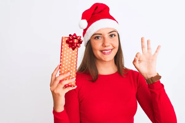 若いです美しい女の子身に着けていますクリスマスサンタ帽子保持ギフト上の隔離された白い背景を行います指でOkサイン 優れたシンボル — ストック写真