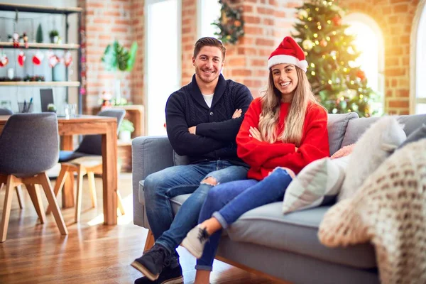 年轻夫妇戴着圣诞老人帽坐在沙发上 围绕着圣诞树 开心的脸上挂着笑容 交叉着双臂看着摄像机 积极的人 — 图库照片