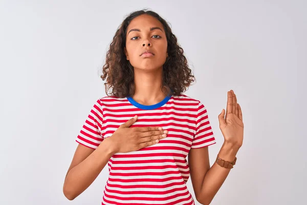 Young brazilian woman wearing red striped t-shirt standing over isolated white background Swearing with hand on chest and open palm, making a loyalty promise oath
