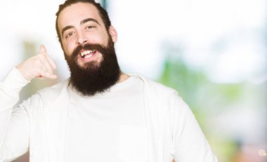 Uzun saçlı ve sakallı genç adam sportif sweatshirt giyen telefonda konuşmak gibi el ve parmakları ile telefon jest yapıyor gülümseyerek. İletişim kavramları.
