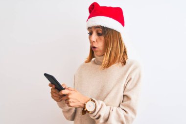 Güzel kızıl saçlı kadın Noel şapkası takıyor ve akıllı telefon kullanıyor. Şoktan korkmuş, korkmuş ve korku ifadesinden heyecanlanmış.