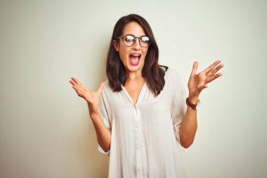 Tişört ve gözlük takan genç güzel bir kadın izole edilmiş beyaz arka planda duruyor gülümsüyor ve kameraya bakıyor iki eliyle ve parmaklarıyla yandan işaret ediyor..