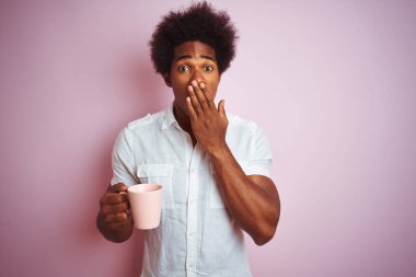 Genç Afro-Amerikalı bir adam bir fincan kahve içerken izole edilmiş pembe arka planda duruyor. Ağzını kapatıyor. Yanlışlıkla utancından ağzı sarsılıyor.