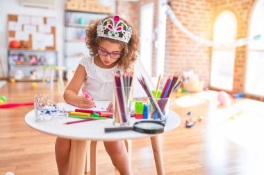 Güzel bebek gözlük takıyor ve prenses tacı takıyor. Anaokulunda kağıt ve keçeli kalemle çiziyor.