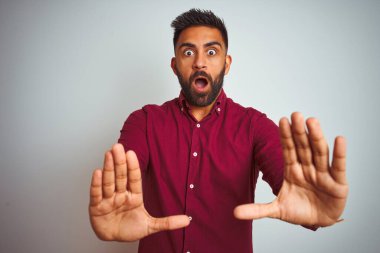 İzole edilmiş gri arka planda duran kırmızı şık tişörtlü genç Hintli adam korku ifadesinden korkuyor ve dehşete kapılıyordu. El hareketlerini durdur, şok içinde bağır. Panik konsepti.