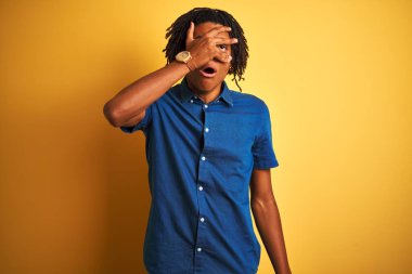 Rastgele kot gömlek giyen Afro bir adam izole edilmiş sarı arka planda dikiliyor. Şok olmuş bir şekilde yüzünü ve gözlerini kapatıyor, utanmış bir ifadeyle parmaklarından bakıyor..