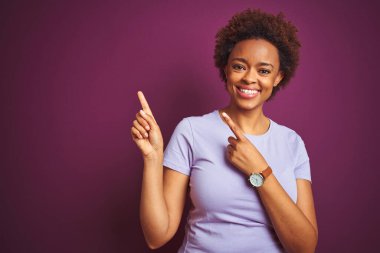 Güzel Afro saçlı, siyah saçlı, güzel bir Afro Amerikalı kadın. Mor arka planda. Gülümsüyor ve kameraya bakıyor. İki eliyle ve parmaklarıyla yan tarafa bakıyor..