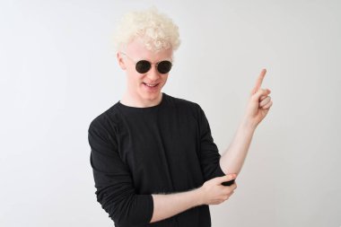 Siyah tişört giyen ve güneş gözlüklü genç albino adam izole edilmiş beyaz arka planda duruyor. Yüzünde kocaman bir gülümseme, eli ve parmağı kameraya bakıyor..