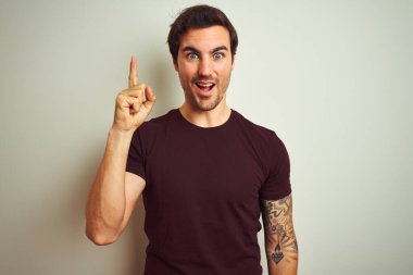 İzole edilmiş beyaz arka plan üzerine mor bir tişört giyen yakışıklı genç adam başarılı bir fikirle işaret ediyor. Heyecanlı ve mutlu. Bir numara..