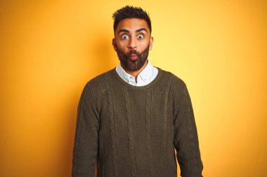 Yeşil kazak ve tişört giyen genç Hintli adam izole edilmiş sarı arka planda dikiliyor dudaklı balık suratı yapıyor, çılgınca ve komik hareketler yapıyor. Komik ifade.