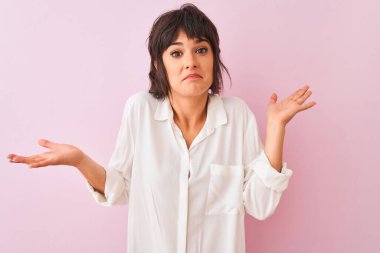 İzole edilmiş pembe arka plan üzerinde duran beyaz gömlekli genç ve güzel bir kadın. Kolları ve elleri havada, kafası karışmış bir ifade. Şüphe kavramı.