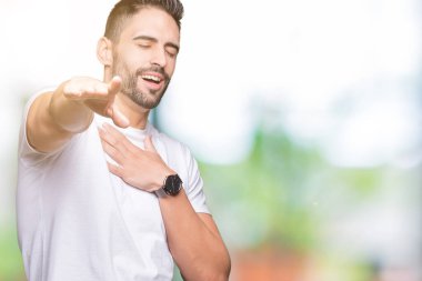 Beyaz tişörtlü yakışıklı adam arka planda sana gülüyor, kamerayı işaret ediyor göğsünde parmak, utanç ifadesi var.