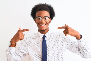 Genç Afro-Amerikalı iş adamı, izole edilmiş beyaz arka planda kravat ve gözlük takıyor, gülümsüyor ve parmaklarıyla ve ağzıyla işaret ediyor. Diş sağlığı konsepti.
