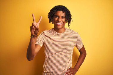 Rasta desenli tişörtlü Afro adam izole edilmiş sarı arka planda durup kameraya bakıp zafer işareti yaparken gülümsüyor. İki numara..