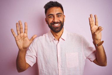 İzole edilmiş pembe arka planda dikilen rahat gömlekli genç Hintli adam sekiz numaralı parmaklarıyla gülümseyerek kendinden emin ve mutlu görünüyor..