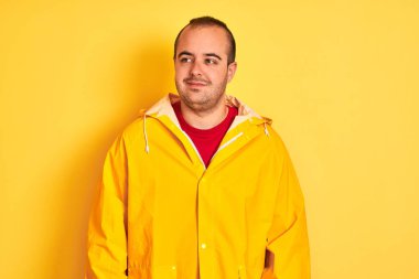 Yağmurluk giyen genç adam izole edilmiş sarı arka planda duruyor gülümsüyor ve yan tarafa bakıyor ve düşünüyor..