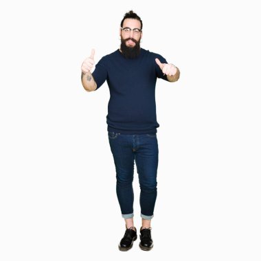 Uzun saç ve sakal gözlük başarı işareti el ile pozitif jest yapıyor giyen genç hipster adamla başparmak gülümseyen ve mutlu. Neşeli bir ifadeyle, kazanan jest kameraya bakıyor.