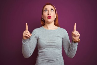 Gri tişört giyen güzel, kızıl saçlı bir kadın ve mor arka plan üzerine çap takmış şaşkın şaşkın yukarı bakıyor ve parmaklarıyla işaret ediyor ve kollarını kaldırıyordu..