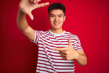 Asyalı, çizgili tişört giyen genç bir Çinli izole edilmiş kırmızı arka planda durup gülümsüyor ve elleri ve parmakları mutlu yüzlü bir çerçeve yapıyor. Yaratıcılık ve fotoğrafçılık kavramı.