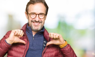 Orta yaş yakışıklı adam gözlük ve kış ceket giydiği yüzünde gülümseme ile kendine güvenen görünümlü, parmaklarıyla gurur ve mutlu kendini işaret.