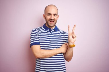 Sakallı genç ve kel bir adam pembe izole edilmiş arka planda günlük mavi tişört giyiyor ve kameraya gülümsüyor zafer işareti yapıyor. İki numara..