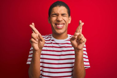 İzole edilmiş kırmızı arka plan üzerinde çizgili tişört giyen genç, yakışıklı Arap adam umut ve gözlerin kapalı bir şekilde gülümseyerek işaret ediyor. Şans ve batıl inanç..