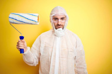 Boyama ekipmanı giyen ve elinde boya rulosu tutan genç bir adam izole edilmiş sarı arka planda, akıllı yüzünde kendine güvenen bir ifadeyle ciddi ciddi düşünüyor.