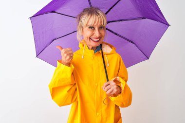 İzole edilmiş beyaz arka plan üzerinde yağmurluk ve mor şemsiye giyen orta yaşlı kadın gülümsüyor.