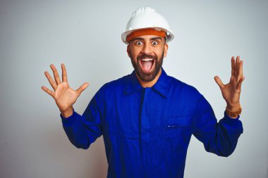 Üniformalı ve kasklı yakışıklı Hintli işçi, izole edilmiş beyaz arka plan üzerinde çılgın ve başarıyı kollarını kaldırıp heyecanla haykırarak kutluyor. Kazanan konsept