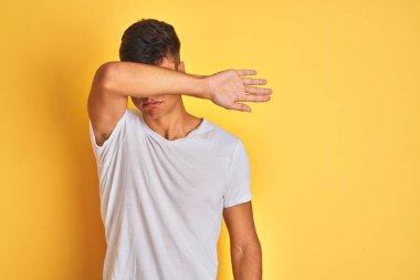 İzole edilmiş sarı arka plan üzerinde duran beyaz tişörtlü genç Hintli adam kollarıyla gözlerini kapatıyor, ciddi ve üzgün görünüyor. Görme, saklama ve reddetme konsepti