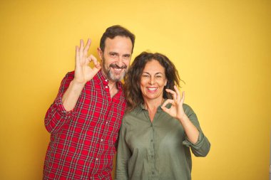 Güzel bir orta yaşlı çift, izole edilmiş sarı arka planda gülümsüyor ve el ve parmaklarıyla 