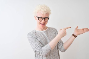 İzole edilmiş beyaz arka plan üzerinde çizgili tişört ve gözlük takan genç albino sarışın adam hayrete düşmüş ve kameraya gülümserken eliyle ve parmağıyla işaret ediyordu..