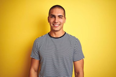 Sarı izole edilmiş arka plan üzerine çizgili tişört giyen genç yakışıklı bir adam kameraya seksi bir ifadeyle bakıyor, neşeli ve mutlu bir yüzle..