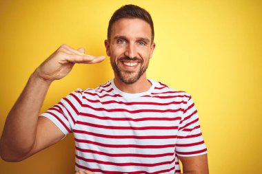 Sarı izole edilmiş arka plan üzerine gündelik kırmızı çizgili tişört giyen genç yakışıklı bir adam. Ellerinde büyük ve büyük beden işareti, ölçü işareti var. Kameraya bakıp gülümsüyor. Ölçüm konsepti.