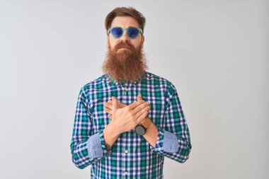 Genç, kızıl saçlı, günlük gömlek ve güneş gözlüğü takan bir adam izole edilmiş beyaz arka planda elleri göğsünde, gözleri kapalı ve yüzünde minnet dolu bir jest ile gülümsüyor. Sağlık kavramı.
