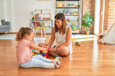 Güzel öğretmen ve sarışın küçük kız kreşteki plastik blokları kullanarak kule inşa ediyorlar.