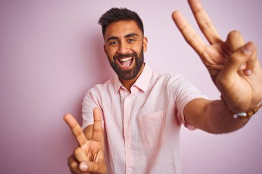 İzole edilmiş pembe arka planda dikilmiş sıradan gömlekler giyen genç Hintli adam kameraya bakıp zafer işareti yaparken gülümsüyor. İki numara..