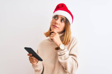 Güzel kızıl saçlı kadın Noel şapkası takıyor ve akıllı telefon kullanıyor. Ciddi bir yüz ifadesi takınıyor. Soru sormayı düşünüyor.