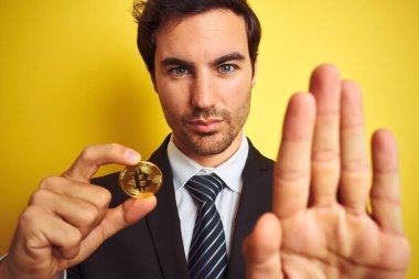 Bitcoin tutan genç yakışıklı işadamı, izole edilmiş sarı arka planda, açık elle, ciddi ve kendinden emin bir ifadeyle dur işareti yapıyor, savunma jesti.