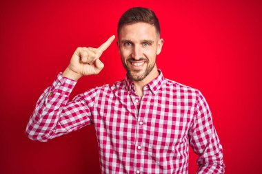 Kırmızı izole edilmiş arka plan üzerinde genç yakışıklı bir adam. Tek parmakla kafasını göstererek gülümsüyor. Harika bir fikir ya da düşünce, iyi bir hafıza.