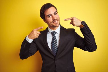 Takım elbiseli ve kravatlı genç yakışıklı işadamı izole edilmiş sarı arka planda gülümseyip parmaklarıyla ve ağzıyla işaret ediyor. Diş sağlığı konsepti.