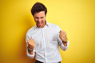 Zarif bir iş adamı, sarı arka plan üzerinde duran zarif gömleğini giyer mutlu ve heyecanlı bir şekilde kollarını kaldırır, gülümser ve başarı için bağırır. Kutlama konsepti.