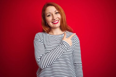 Genç kızıl saçlı kadın, lacivert gömlek giyiyor. Kırmızı izole edilmiş arka plan üzerinde neşeli duruyor. Yüzünde mutlu ve doğal bir ifadeyle, elleri ve parmaklarıyla yüzünü gösteren bir gülümseme var.