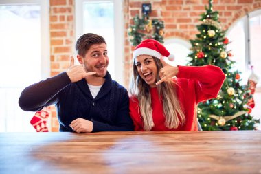 Noel Baba şapkası takan genç bir çift evde Noel ağacının etrafında oturmuş gülümsüyor telefonla el ve parmaklarıyla telefonla konuşuyor gibi yapıyor. İletişim kavramları.