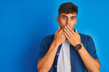 İzole edilmiş mavi arka planda duran gömlek giyen genç Hintli adam yanlışlıkla elleriyle ağzını kapatırken şok oldu. Gizli konsept.