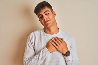 İzole edilmiş beyaz arka plan üzerinde duran süveter giyen genç Hintli adam elleri göğsünde, gözleri kapalı ve yüzünde minnet dolu bir jest ile gülümsüyor. Sağlık kavramı.