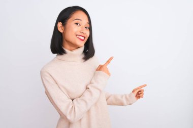 Balıkçı yaka kazak giyen genç bir kadın izole edilmiş beyaz arka plan üzerinde duruyor gülümsüyor ve kameraya bakıyor iki eliyle ve parmaklarıyla yan tarafı işaret ediyor..