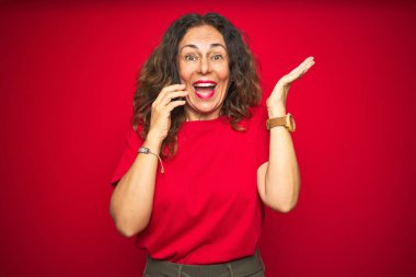 Orta yaşlı bir kadın telefonda kırmızı izole edilmiş arka planda konuşuyor çok mutlu ve heyecanlı, kazanan yüz ifadesi zaferi büyük bir gülümsemeyle kutluyor ve ellerini kaldırıyor.