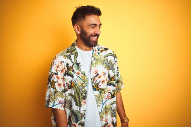 Tatilde olan genç Hintli bir adam yaz çiçekli gömleğini giyip izole edilmiş sarı arka planda, yüzünde gülümseme, doğal bir ifade ile yan yana bakıyor. Kendinden emin gülen.